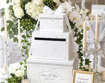 May include: A three-tiered white card box set with rhinestone accents and a white bow, displayed on a table with a floral arrangement and candles. The bottom box has the text "Jessica & Thad, October 4, 2016". A framed sign reads "Cards and Well Wishes for Jessica & Thad."