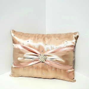 May include: A decorative pillow with a blush pink satin bow and a pearl-like button. The pillow is covered in a champagne-colored satin fabric and has small rhinestones scattered across the surface.