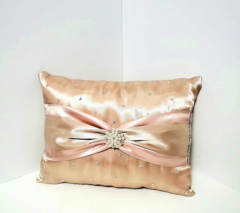 May include: A decorative pillow with a blush pink satin bow and a pearl-like button. The pillow is covered in a champagne-colored satin fabric and has small rhinestones scattered across the surface.