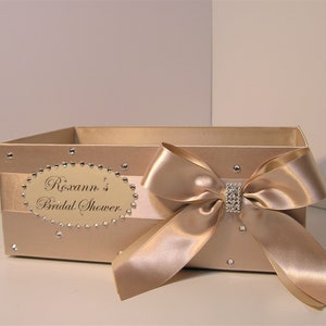 May include: A beige satin box with a large beige satin bow and a rhinestone embellishment. The box has a label that reads "Roxann's Bridal Shower".