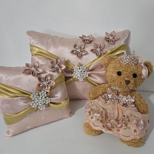 May include: A set of three decorative pillows with pink satin fabric, gold ribbon, and jeweled accents. The pillows feature a floral design and are paired with a plush teddy bear wearing a pink dress and a jeweled crown.