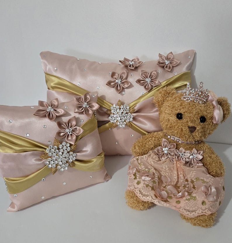 May include: A set of three decorative pillows with pink satin fabric, gold ribbon, and jeweled accents. The pillows feature a floral design and are paired with a plush teddy bear wearing a pink dress and a jeweled crown.