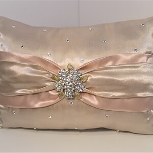 May include: A decorative pillow with a blush pink satin bow and a rhinestone brooch. The pillow is covered in a champagne-colored satin fabric and has scattered rhinestones.