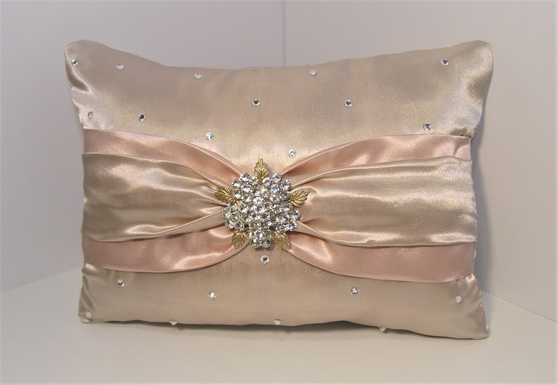 May include: A decorative pillow with a blush pink satin bow and a rhinestone brooch. The pillow is covered in a champagne-colored satin fabric and has scattered rhinestones.