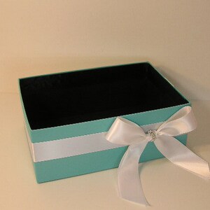 May include: A turquoise gift box with a white satin ribbon and a rhinestone bow. The box is empty and ready to be filled with a special gift.