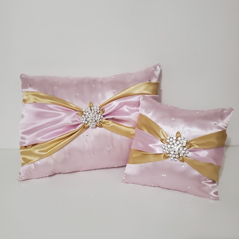 May include: Two decorative pillows with pink satin fabric and gold satin ribbon accents. Each pillow features a large rhinestone brooch in the center.