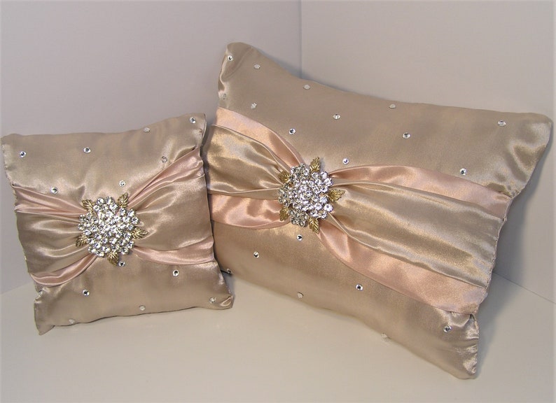 May include: Two decorative pillows with a satin finish. The larger pillow is beige with a pink satin bow and a jeweled brooch. The smaller pillow is beige with a pink satin bow and a jeweled brooch.