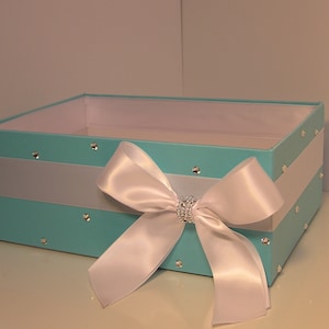 May include: A turquoise gift box with a white satin ribbon and bow. The box is decorated with silver rhinestones.