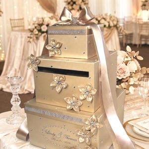 May include: A three-tiered gold wedding card box with floral embellishments and a large bow. The box has a slot for cards and is decorated with rhinestones and satin ribbons. The box has the names "Lindy & Thomas" and the date "Mar 31, 2018" on it.