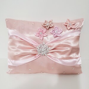 May include: A pink satin pillow with a large bow and a rhinestone embellishment. The pillow is decorated with four pink satin flowers.