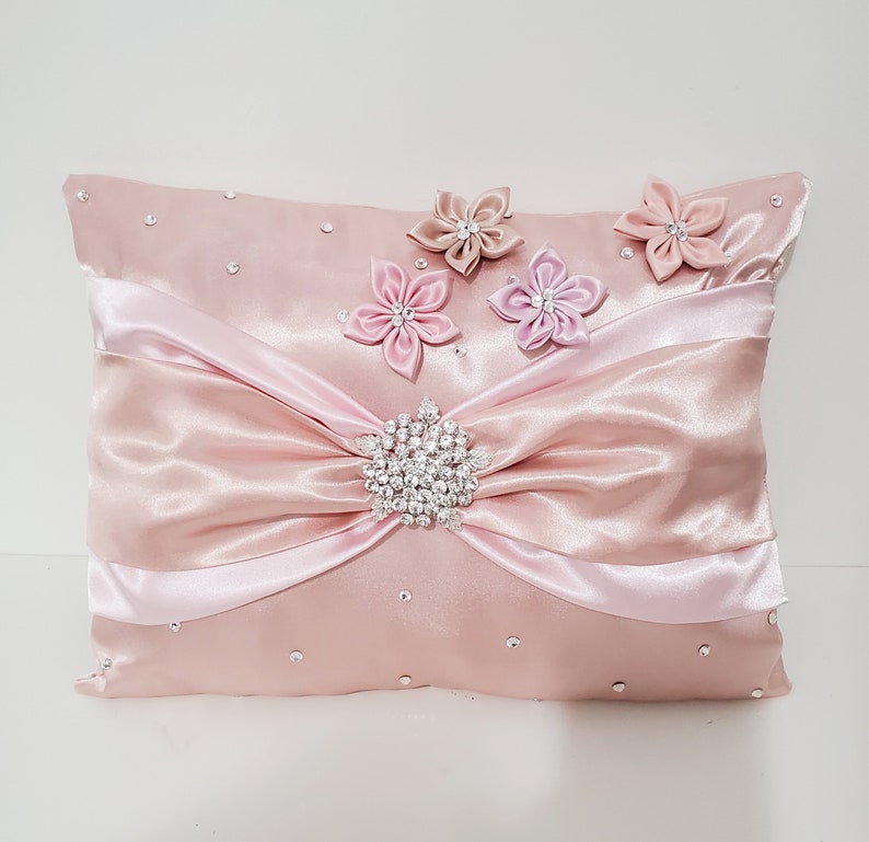May include: A pink satin pillow with a large bow and a rhinestone embellishment. The pillow is decorated with four pink satin flowers.