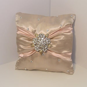 May include: A blush pink satin pillow with a large, gold-toned rhinestone brooch in the center. The pillow is decorated with small rhinestones.