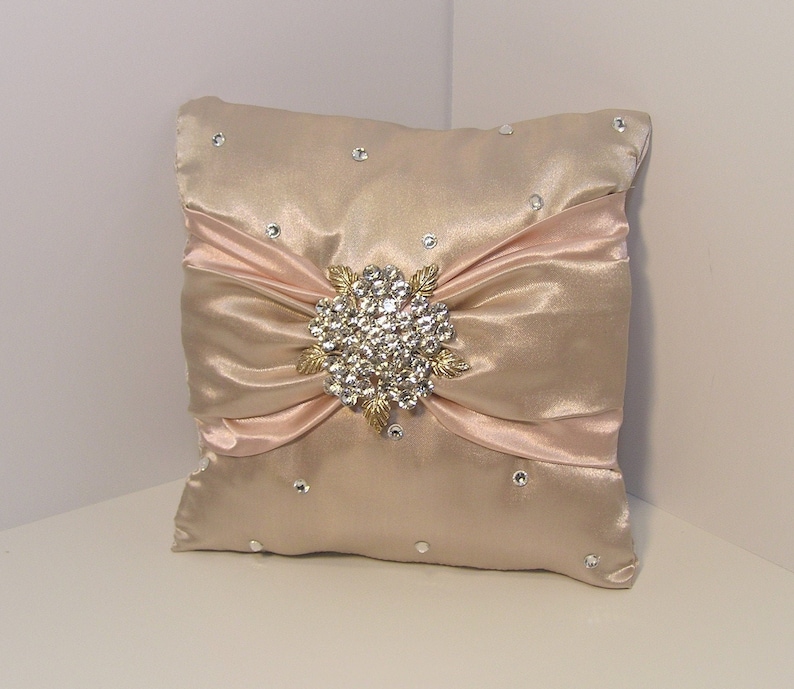 May include: A blush pink satin pillow with a large, gold-toned rhinestone brooch in the center. The pillow is decorated with small rhinestones.