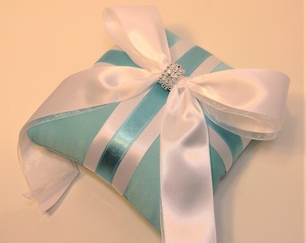 May include: A light blue ring pillow with white satin ribbon and a rhinestone embellishment. The pillow is decorated with white and light blue satin stripes.