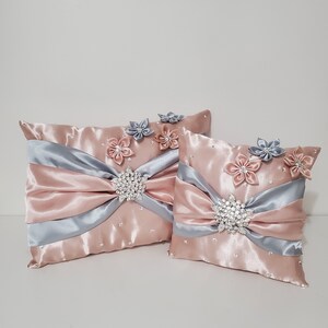 May include: Two decorative throw pillows with a blush pink satin fabric and silver satin ribbon accents. The pillows are embellished with silver and blush pink fabric flowers and jeweled brooches.