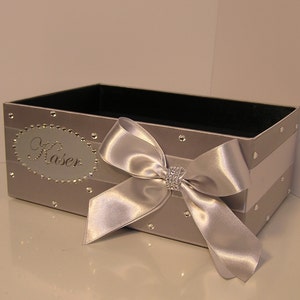 May include: A silver gift box with a white satin ribbon and bow. The box is decorated with rhinestones and has the name "Kaser" written on it.