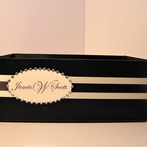 May include: Black box with white stripes and a white oval label with the text "Jamie W Scott" in black script. The oval label is surrounded by silver studs.