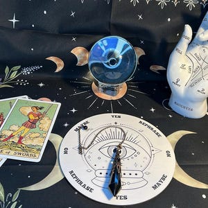 Könnte beinhalten: Eine Sammlung von Wahrsage-Werkzeugen auf einem schwarzen Tuch mit Himmels- und Blumenmustern. Enthält Tarotkarten, eine Kristallkugel, eine Handlesehand, ein Pendelbrett und ein Kristallprisma. Die Tarotkarten zeigen Illustrationen und Text.