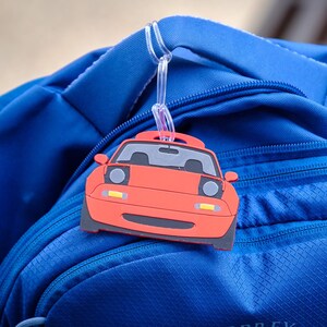 May include: A red car-shaped luggage tag with black and gray details hangs from a blue backpack. The tag features headlights, a windshield, and a license plate area. The backpack has multiple zippers and a textured surface.