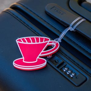 Op de afbeelding: Een rode en witte koffiedruppel bagagelabel bevestigd aan een zwarte koffer. Het label is een gestileerde illustratie van een koffiedruppelaar. De koffer heeft een combinatieslot en een zwarte handgreep.