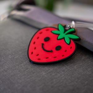 May include: A red strawberry-shaped luggage tag with a smiling face, black seeds, and green leaves. The tag has a black outline and a clear plastic loop for attachment. The background is a grey surface.