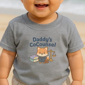 May include: A gray short-sleeved t-shirt with the text "Daddy's CoCounsel" in blue. The shirt features a cartoon fox wearing a suit, holding a briefcase, and standing next to a scale and books. The shirt is for babies and toddlers.
