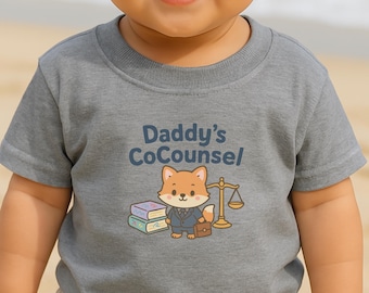 May include: A gray short-sleeved t-shirt with the text "Daddy's CoCounsel" in blue. The shirt features a cartoon fox wearing a suit, holding a briefcase, and standing next to a scale and books. The shirt is for babies and toddlers.