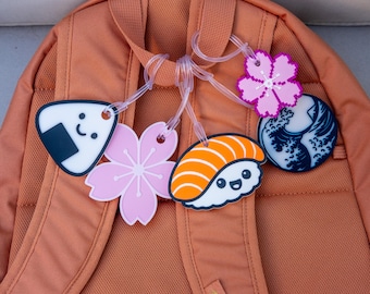 May include: A collection of colorful, Japanese-themed luggage tags attached to a backpack. The tags feature designs of sushi, cherry blossoms, a rice ball, and a wave pattern. The tags are made of a flexible material and are connected by clear plastic loops.