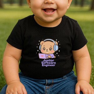 May include: Black t-shirt with a cartoon bear wearing headphones and using a laptop. The text "Junior Software Engineer" is below the bear. The child is wearing blue jeans and is smiling.