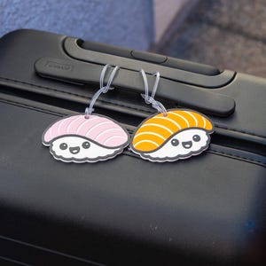 May include: Two sushi-shaped luggage tags with clear plastic loops hang from a black suitcase handle. One tag features pink "fish" and the other orange "fish" atop white rice, both with smiling faces and black outlines.