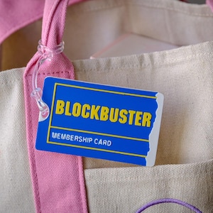 Op de afbeelding: Een beige canvas draagtas met roze handvatten en randen. Een blauwe Blockbuster-lidmaatschapskaart met gele tekst is bevestigd. De tas heeft een zak.