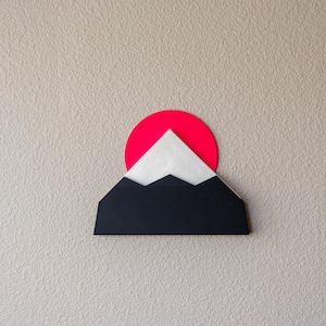 Può includere: Un portatovaglioli a forma di montagna nera con un sole rosso e tovaglioli bianchi. Il supporto è montato su una parete beige. Il design è una rappresentazione minimalista di un paesaggio montano.