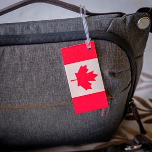 May include: A gray travel bag with a red and white Canadian flag luggage tag attached. The tag features a red maple leaf on a white background, with red stripes at the top and bottom. The bag has a brown handle and a zipper.