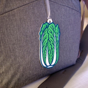 Puede incluir: Una etiqueta de equipaje en forma de col china verde y blanca con un contorno azul cuelga de una mochila gris. La etiqueta está hecha de un material duradero y presenta una ilustración detallada del vegetal.