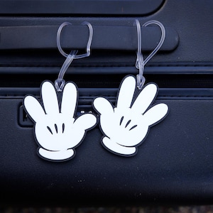 May include: Two white cartoon hand luggage tags with black outlines and black details hang from a black suitcase. The tags are made of a durable material and are designed to be easily attached to luggage.