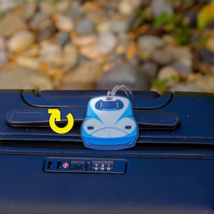 May include: A blue and white train-shaped luggage tag is attached to a black suitcase. The tag has a clear loop for attachment. The suitcase has a combination lock and a handle. The background is blurred with rocks and foliage.