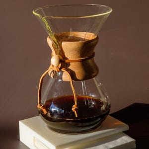 Può includere: Una caffettiera in vetro trasparente con un collare in legno e un laccio in pelle, riempita di caffè scuro. Si trova su una pila di filtri di carta bianchi, ideale per la preparazione del caffè.