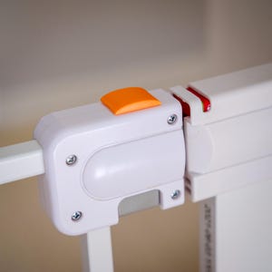 May include: Close-up of a white baby gate with an orange release button. The gate is made of white plastic and metal, with visible screws and a locking mechanism. The gate is designed to secure a doorway or opening.