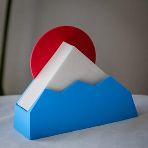 Può includere: Un portatovaglioli blu a forma di montagna, contenente tovaglioli bianchi. Un disco rosso è posto dietro i tovaglioli, simile al sole. Il supporto è realizzato in un materiale solido.