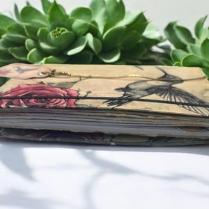 May include: A closed journal with a floral and hummingbird design. The cover is beige with a red rose and a black and white hummingbird illustration. The journal is bound with a black elastic band.