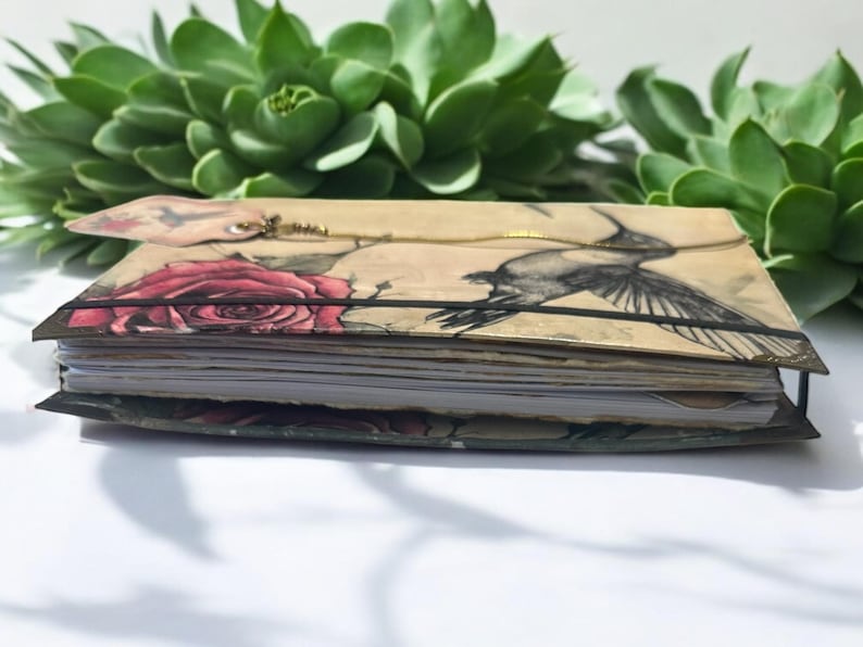 May include: A closed journal with a floral and hummingbird design. The cover is beige with a red rose and a black and white hummingbird illustration. The journal is bound with a black elastic band.