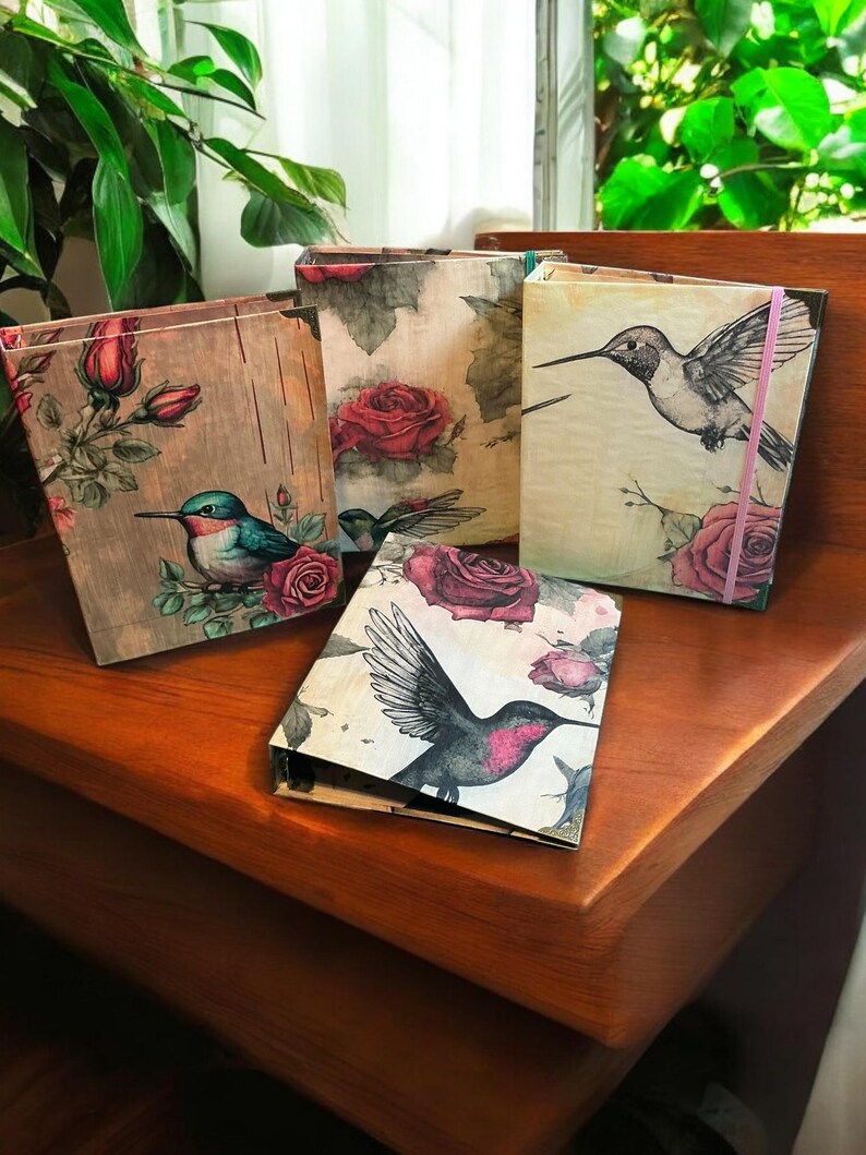 May include: Four decorative journals with floral and hummingbird designs. The journals have a rustic, vintage aesthetic and feature a variety of colors, including pink, green, blue, and brown.