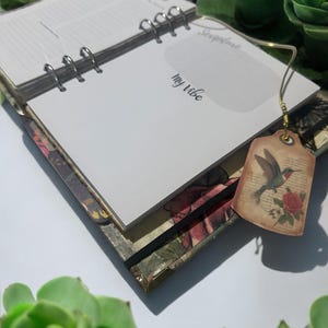 May include: A white journal with a floral design on the cover. The journal has a ring binding and a bookmark with a hummingbird illustration. The text "My Vibe" is written on the cover.