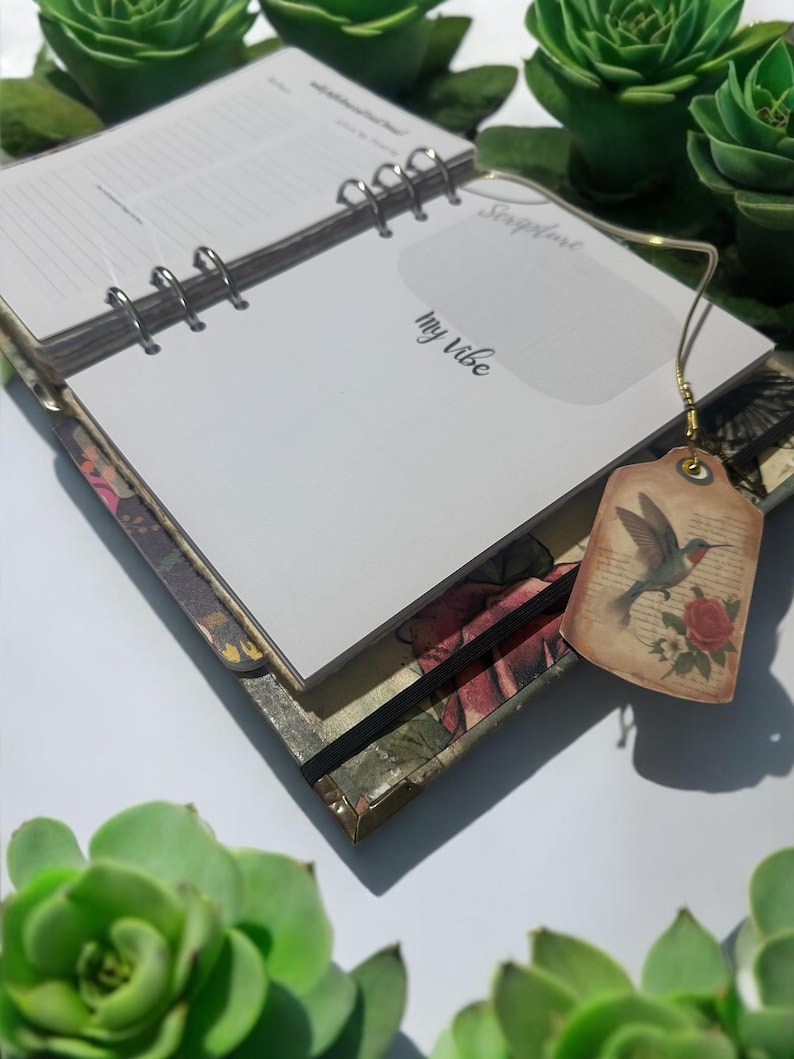 May include: A white journal with a floral design on the cover. The journal has a ring binding and a bookmark with a hummingbird illustration. The text "My Vibe" is written on the cover.