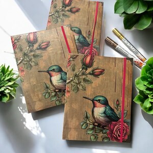 May include: Three brown journals with a floral design featuring hummingbirds and roses. The journals have red ribbon closures.