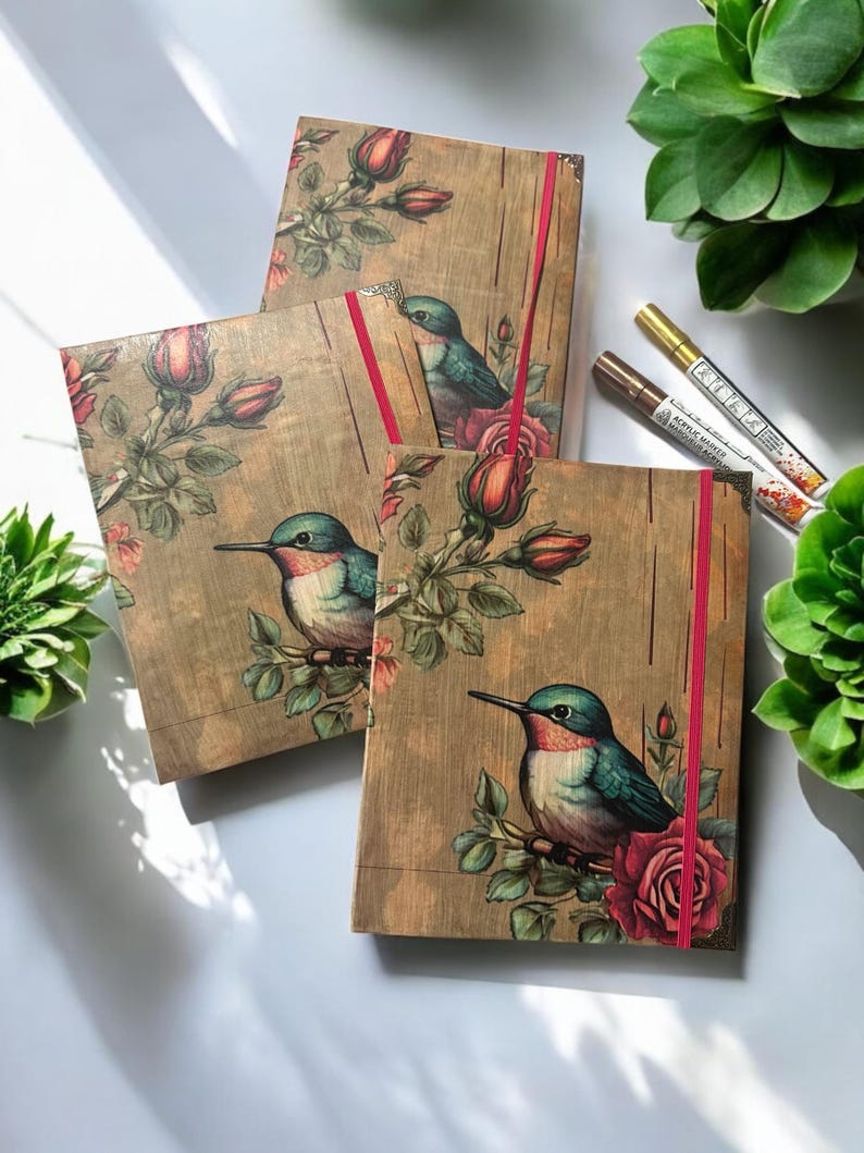 May include: Three brown journals with a floral design featuring hummingbirds and roses. The journals have red ribbon closures.