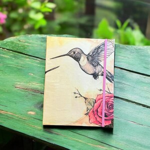 May include: A closed journal with a hummingbird and a rose on the cover. The journal has a pink ribbon bookmark and gold accents.