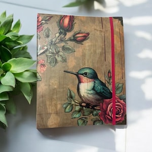 May include: A brown notebook with a floral design and a hummingbird perched on a branch with a red rose. The notebook has a red elastic closure.