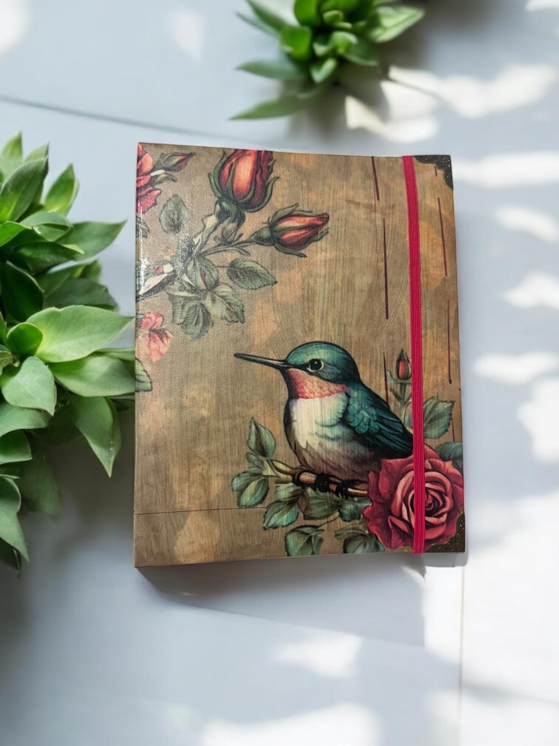 May include: A brown notebook with a floral design and a hummingbird perched on a branch with a red rose. The notebook has a red elastic closure.