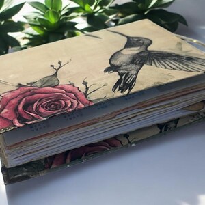 May include: A handmade journal with a beige cover featuring a watercolor illustration of a hummingbird and a rose. The journal has a gold-colored spine and is bound with multiple pages.